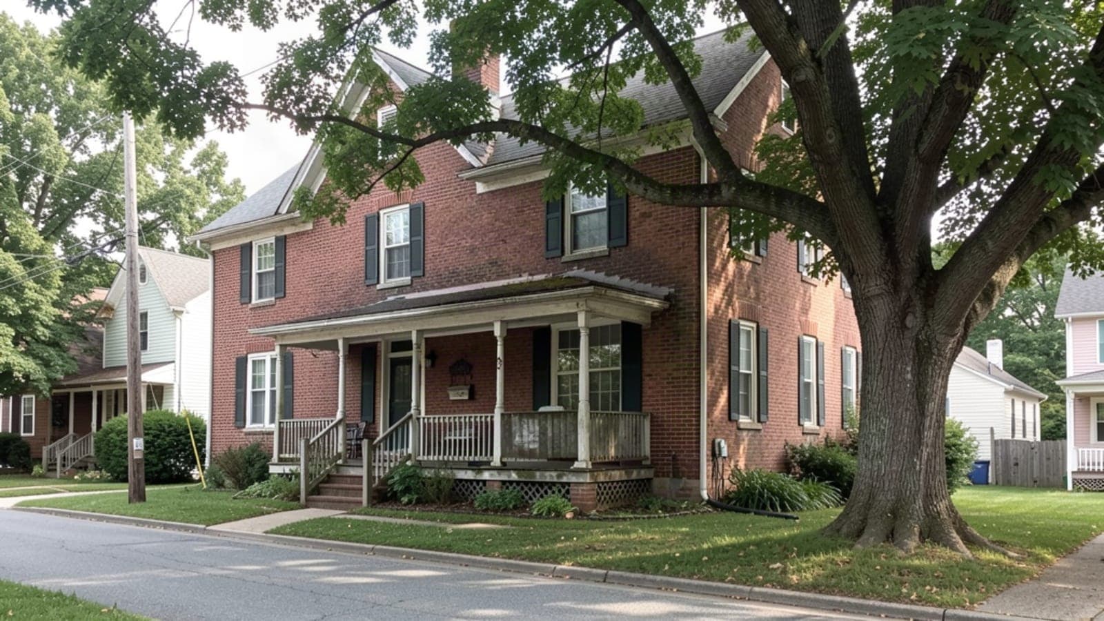 Older brick house with a front porch and a large shade tree on a quiet street