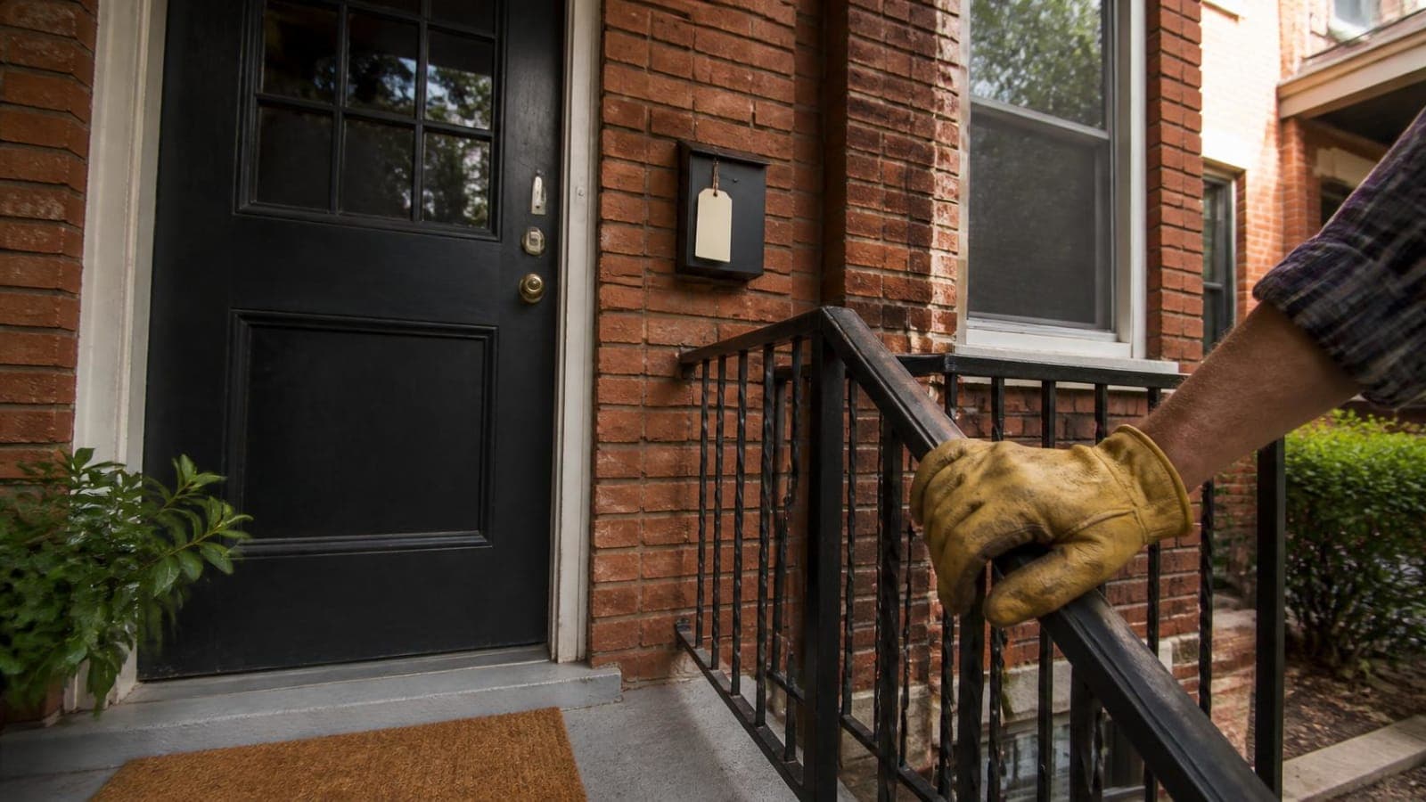 Gloved hand on a porch railing in front of a dark front door on a brick home