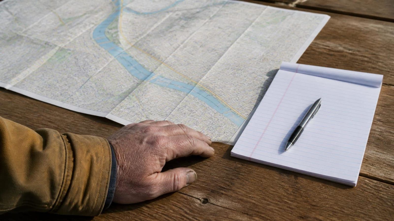 Hand resting on a paper map next to a notepad and pen on a wooden table