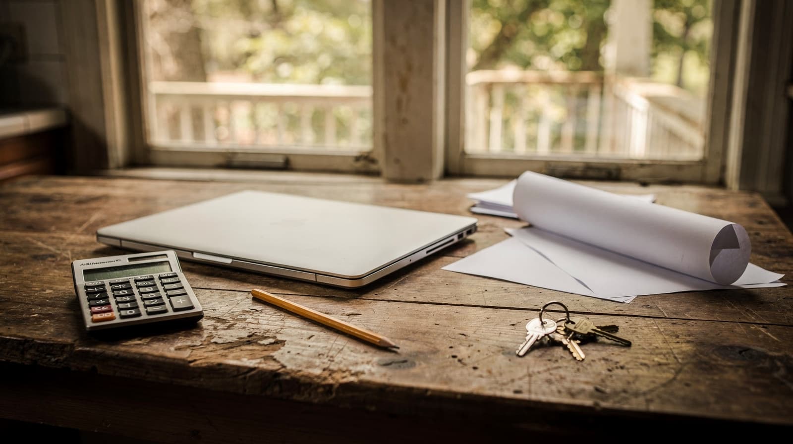 Wooden kitchen table with a closed laptop, calculator, pencil, house keys and blank papers by a porch window