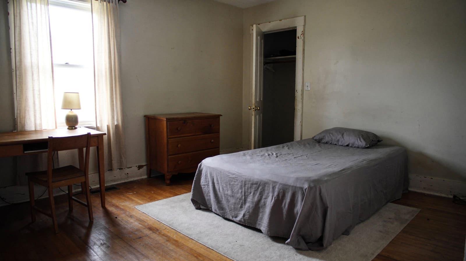 Simple bedroom with a made bed, wooden desk and chair, dresser, lamp, open closet and a curtained window